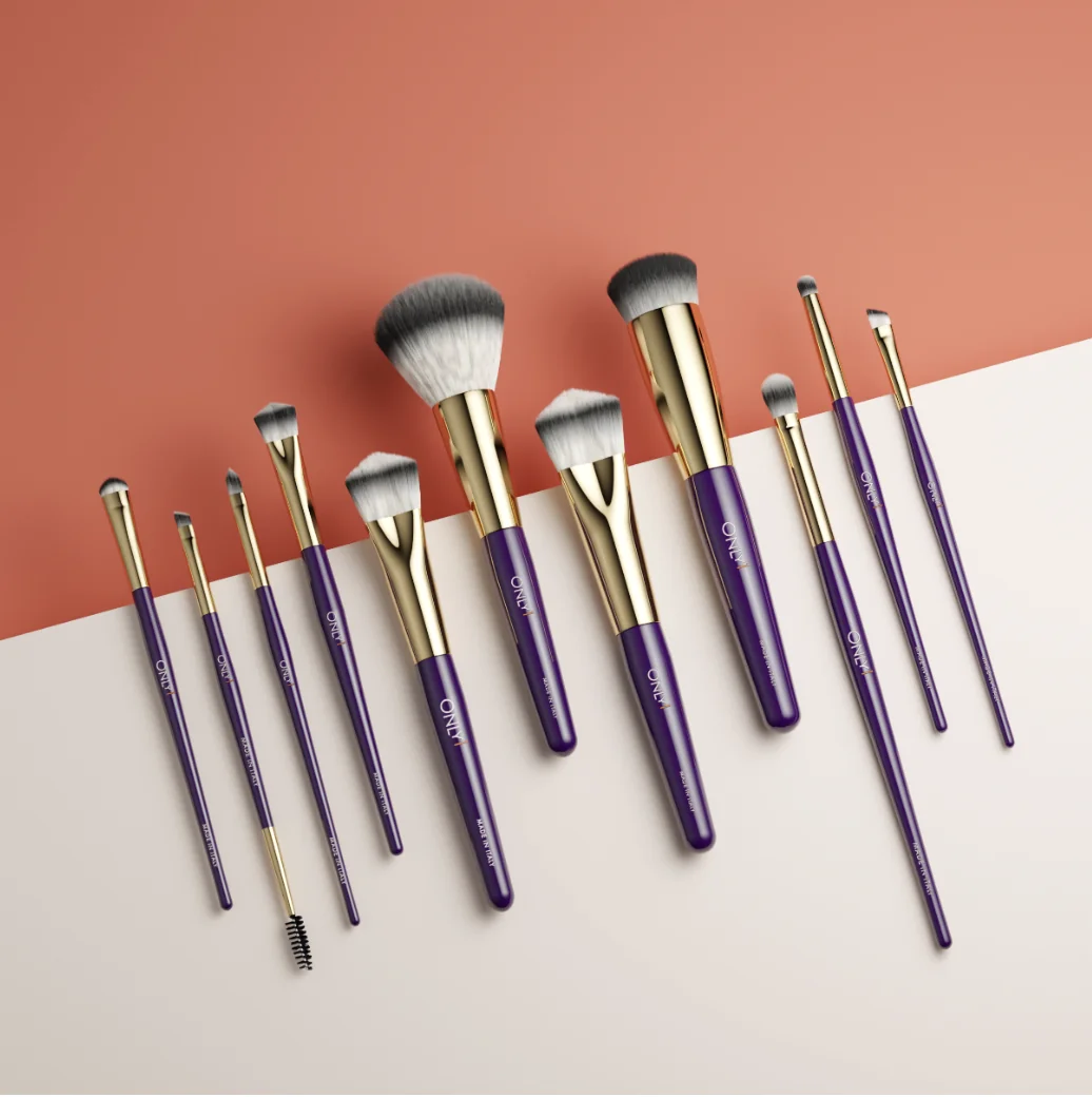 Set of makeup brushes with purple handles and gold accents, arranged on a two-toned peach and white surface. The set includes various sizes and types of brushes.