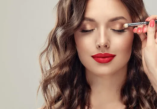 With a bold red lip ,a woman applies shimmery bronze eyeshadow, showcasing one of the best eyeshadow colors for olive skin.