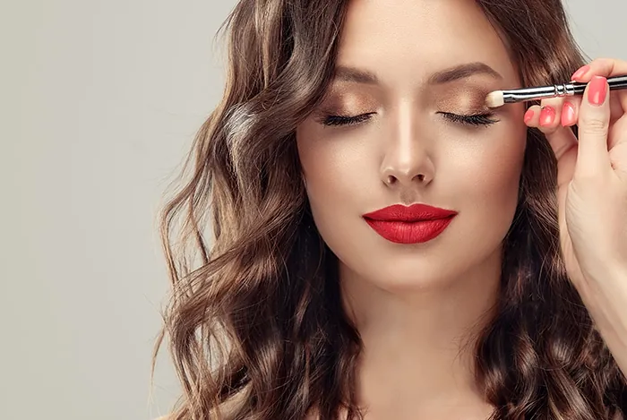 With a bold red lip ,a woman applies shimmery bronze eyeshadow, showcasing one of the best eyeshadow colors for olive skin.